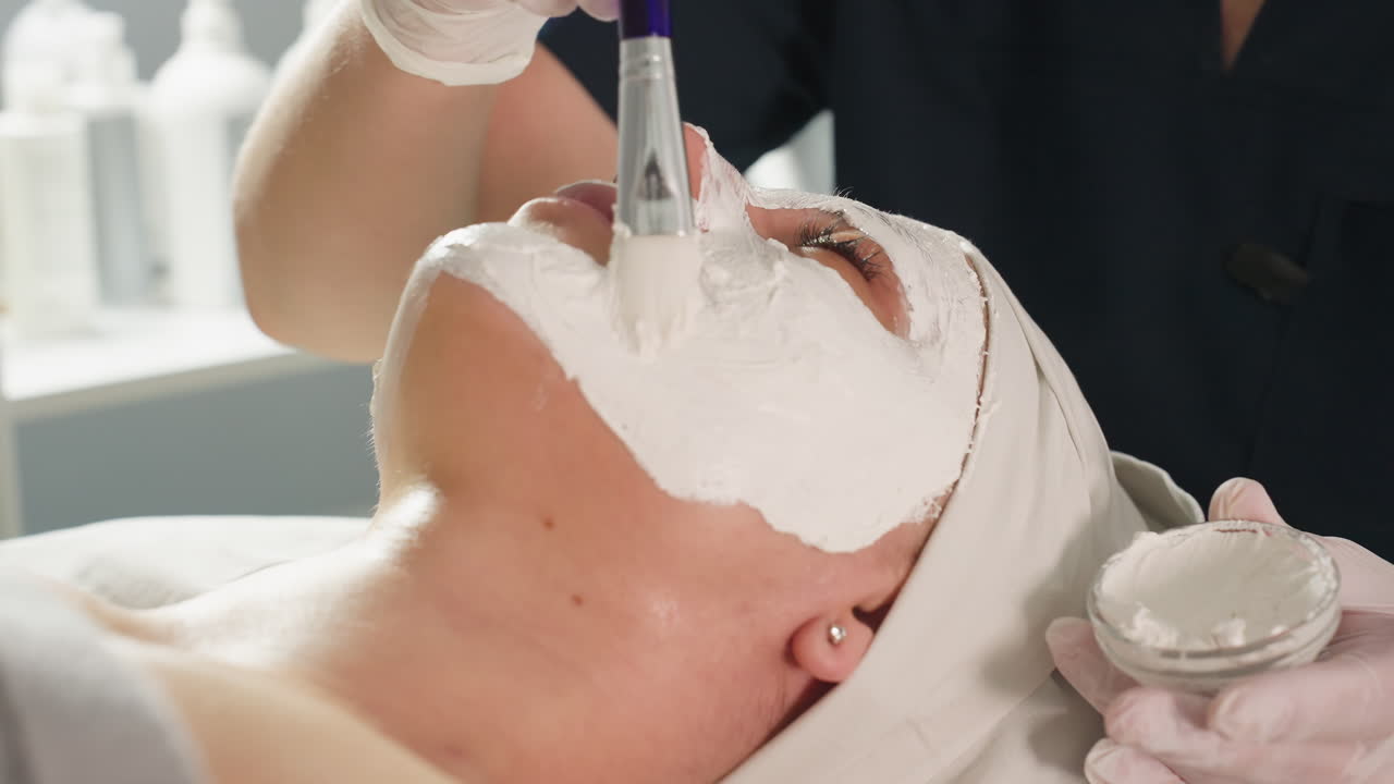 Beauty expert applying thick face mask carefully with soft silicone brush on face of customer during relaxing spa treatment under bright light showcasing professional skincare technique and precision