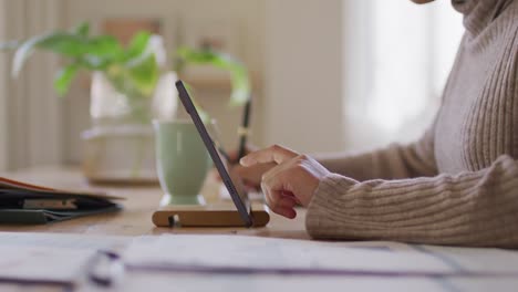 Video-of-midsection-of-biracial-woman-sitting-at-desk-at-home-working-on-tablet