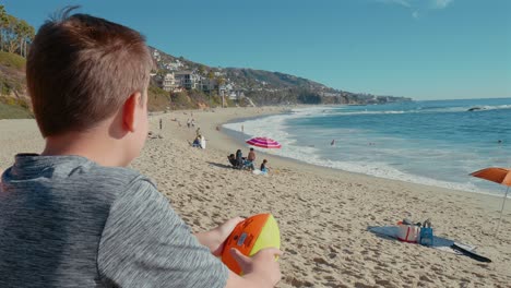 Niño-Observando-Las-Olas-Rompiendo-En-La-Playa,-Sosteniendo-Una-Pelota-De-Fútbol