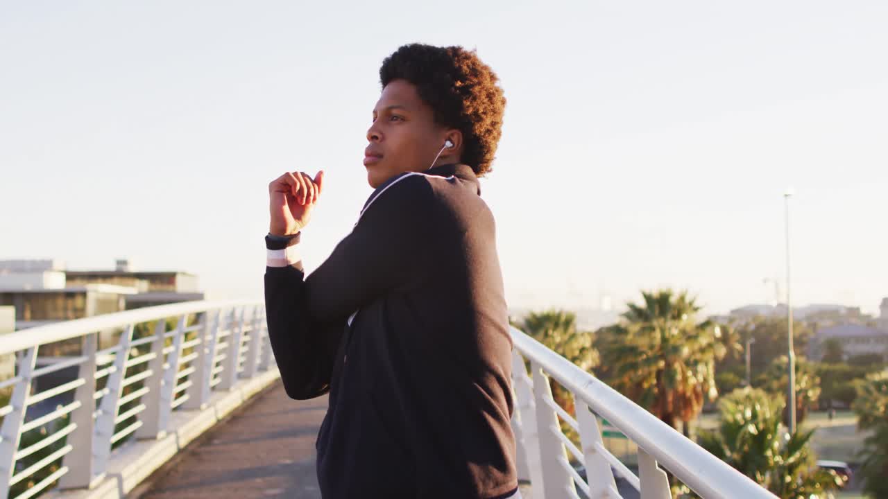 African american man wearing earphones exercising outdoors, standing on footbridge stretching arms