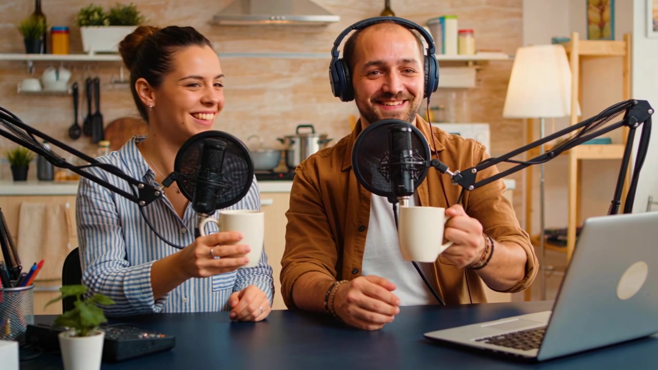 Microfoni in diretta, co-conduttori con giacca marrone e maglia a righe che tengono tazze al tavolo della cucina per un podcast.