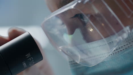 вђ‹вђ‹lab scientist in protective mask and goggles looking through microscope