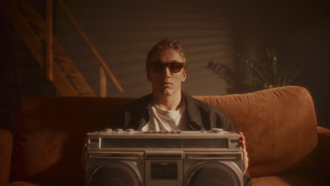 portrait of young man in sunglasses sitting on sofa, holding retro boombox