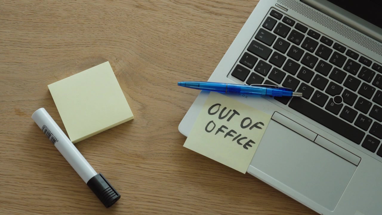 A man places an "Out of Office" sticky note on his laptop, symbolizing vacation, leave, remote work status, and temporary unavailability.