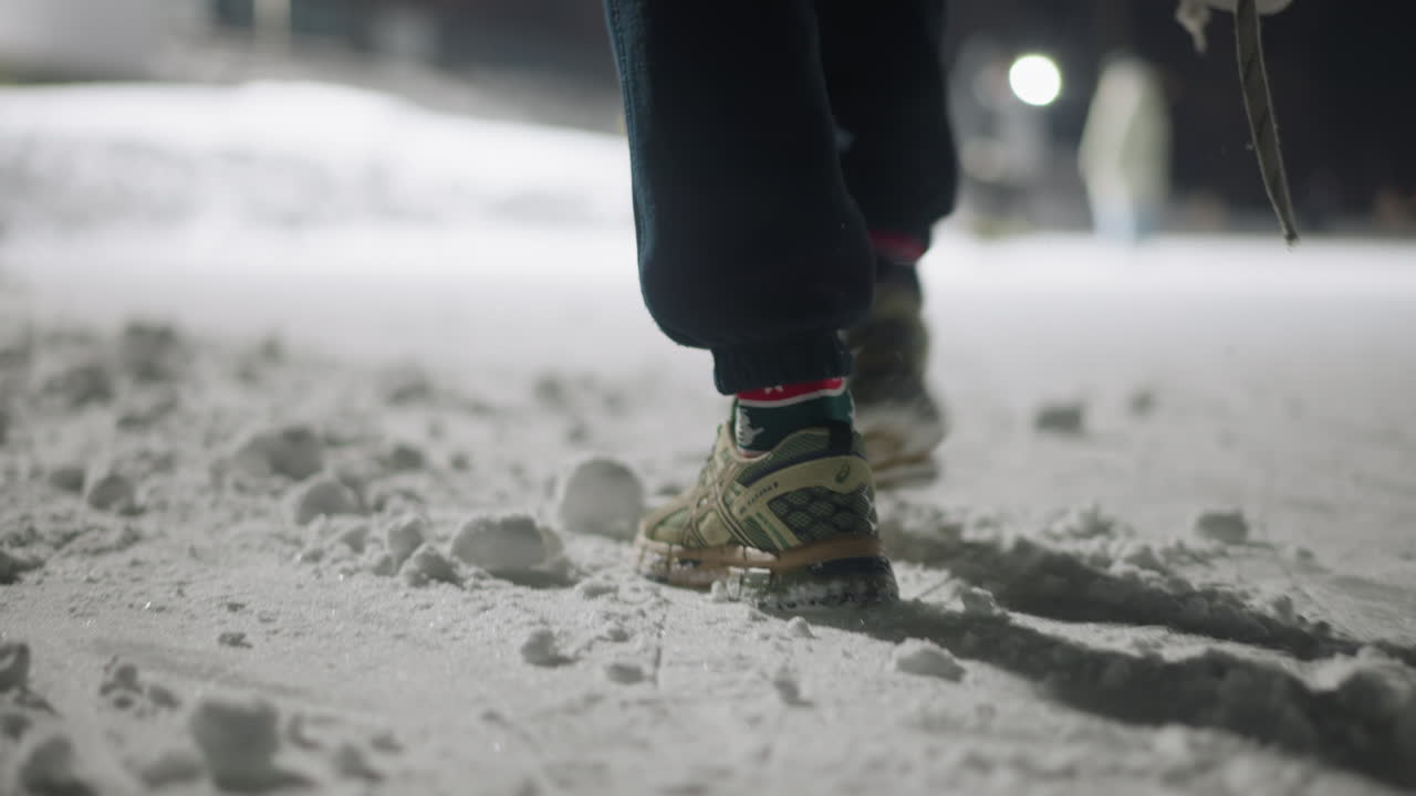 Rear view of person walking on compact snow surface outdoors at night, wearing sneakers and socks, casting long shadow across icy ground with footprints marking path in cold winter setting