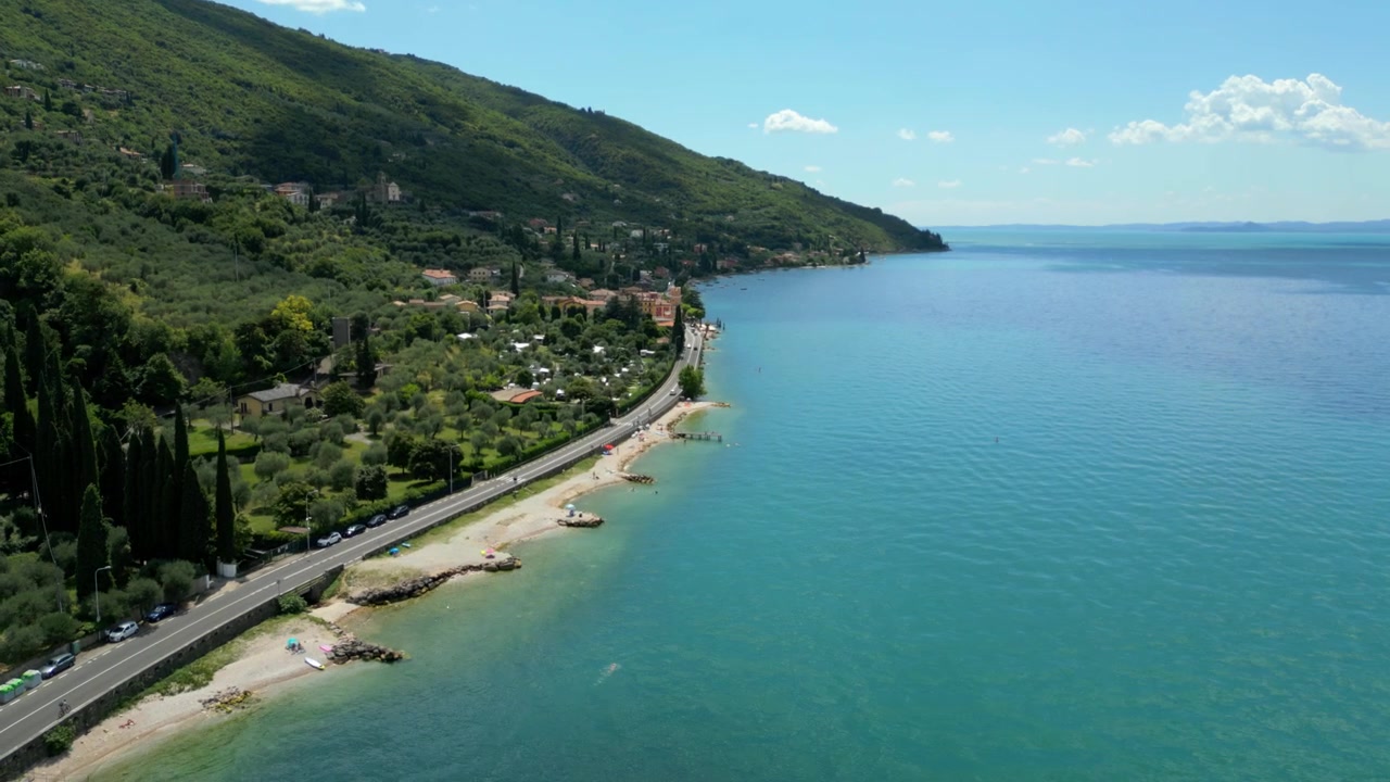 Vista aerea di una strada costiera che costeggia un lago blu e un villaggio collinare in una giornata di sole.