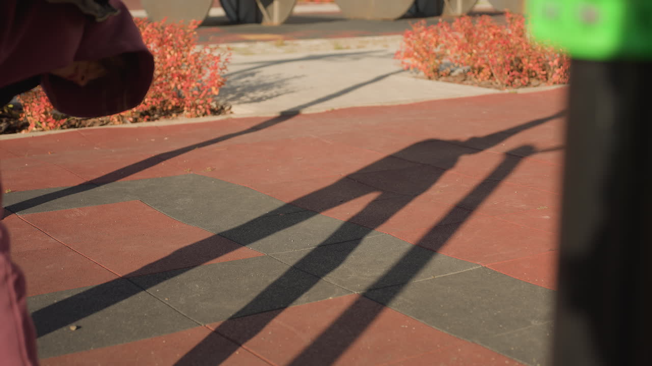 Shadow of gym enthusiast bending down to touch toes on outdoor gym court tiles under bright sunlight, long stretch silhouette cast across patterned rubber surface with red shrub border
