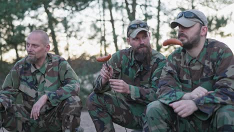 hombres militares en uniformes moro descansando después de un turno de la tarde en la base en el campo sentados en cajas afuera preparando una fogata recogiendo salchichas en palos hombre con barba corta carne con un cuchillo