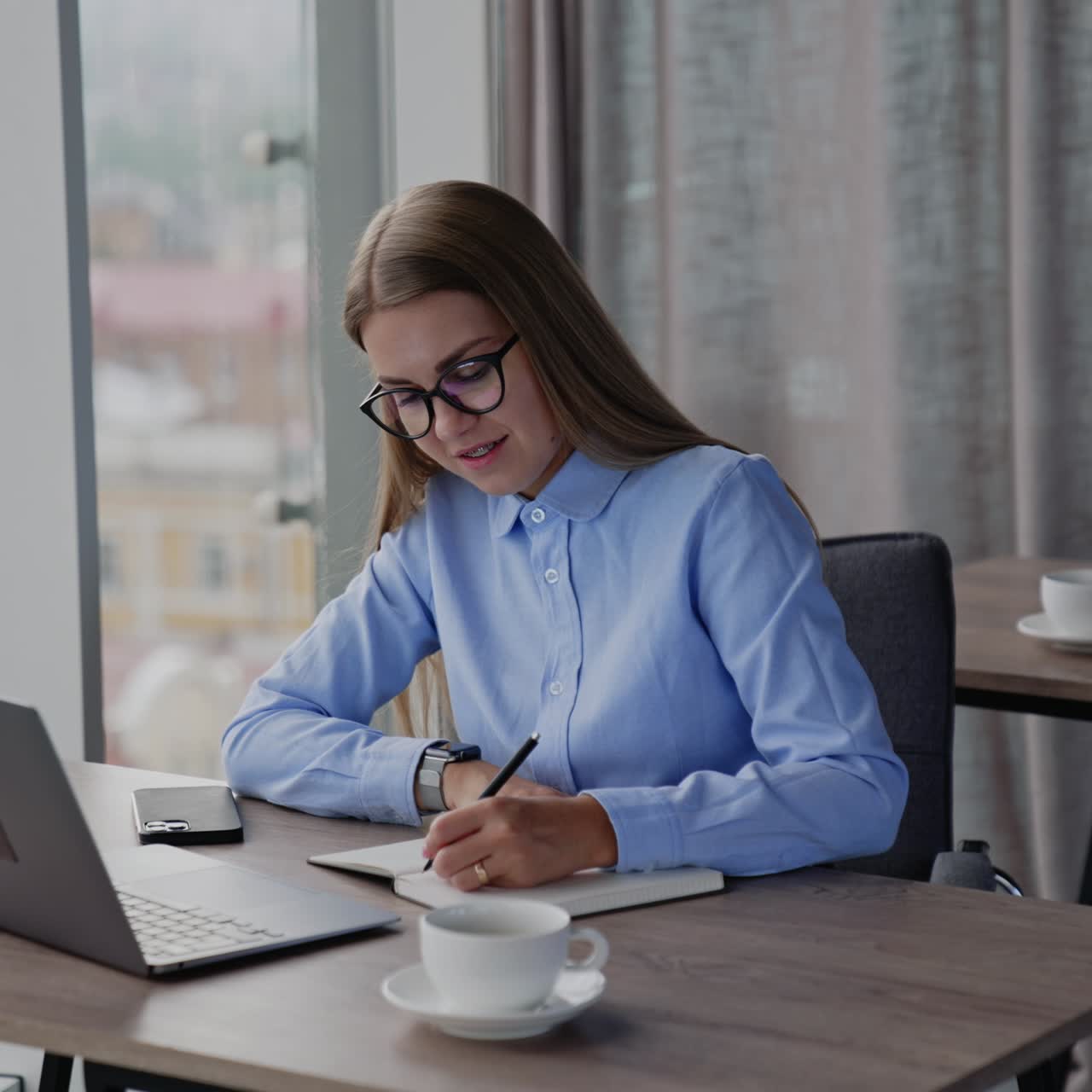 Signora che lavora in ufficio al computer. Donna dai capelli lunghi che prende appunti su un quaderno e poi beve un caffè, guardando fuori dalla finestra