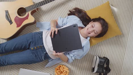 top view of asian teen girl with snacks using tablet while lying on carpet on the floor at home