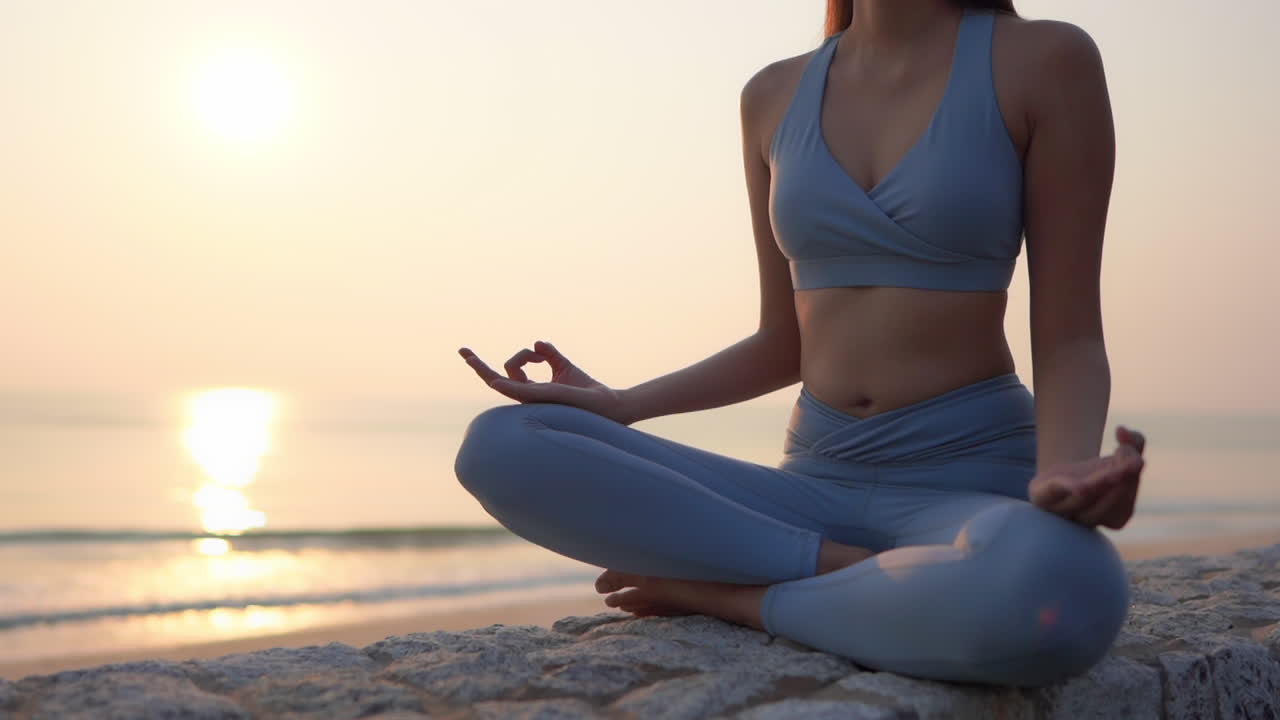 Slim female meditates in lotus pose on stone wall near sea at golden sunset