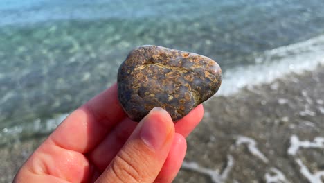 Hand-holding-and-turning-a-beautiful-smooth-grey-rock-with-yellow-dots-at-the-beach-with-turquoise-sea-water-and-waves-in-Manilva-Spain,-sunny-summer-day,-4K-static-shot