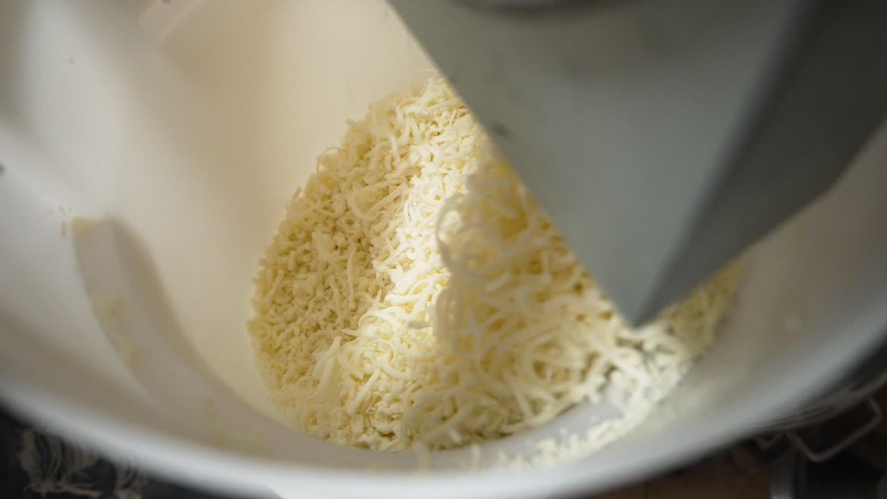 Grattugia elettrica industriale per formaggio lavora sfilacciati di mozzarella in un secchio commerciale bianco per una pizzeria, ripresa ravvicinata dall'alto in slow motion 4K.