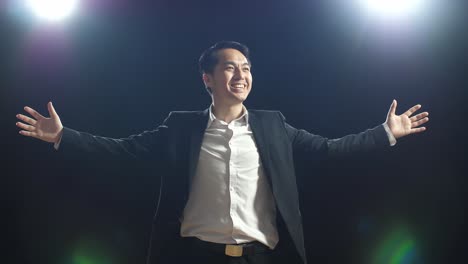 smiling asian speaker man in business suit holds his hands together and spreads out his arms while speaking in the black screen studio