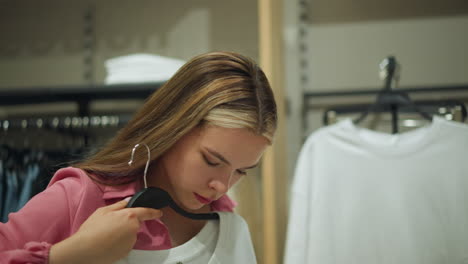 young lady wearing unbuttoned pink top retrieves white top from a rack of other white tops, thoughtfully evaluating it close to her body, other clothes are well arranged in the background