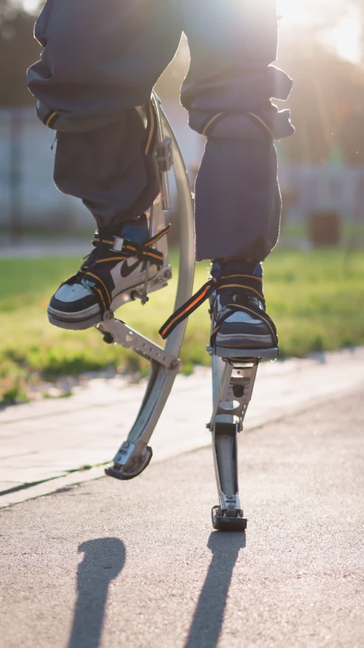 Person walking on spring stilts outdoors on paved park path, wearing dark pants and white sneakers, captured from slightly low angle. Active movement and bouncing