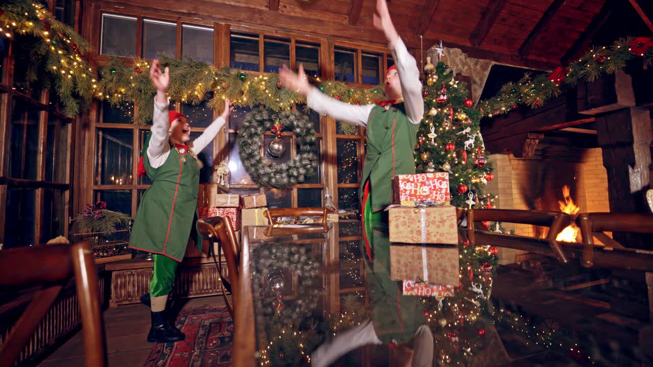 Two elves are standing in specially decorated room near the table and throwing presents to each other. Feast fireplace in the background. Christmas spirit concept close up video. Christmas eve. New Year is coming. Elves are putting presents on the table.