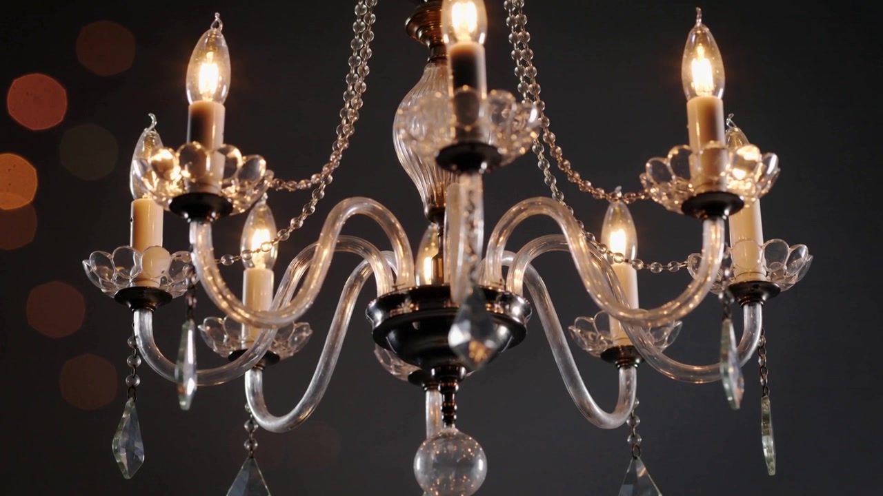 La telecamera zooma verso il basso, evidenziando un lampadario di cristallo con lampadine a forma di candela e cristalli a goccia, in studio.