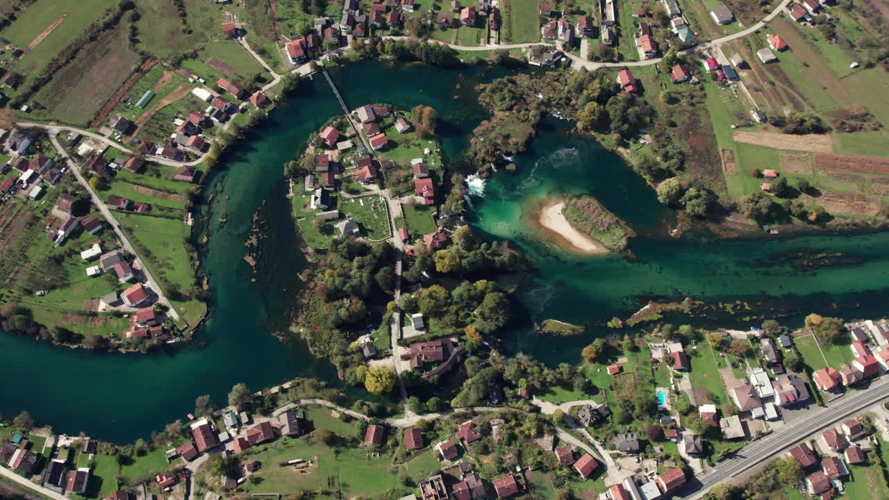 Top-down drone footage of the Una River curving around a small island near Bihac, Bosnia and Herzegovina. Clear turquoise water, houses, and green fields captured in bright daylight