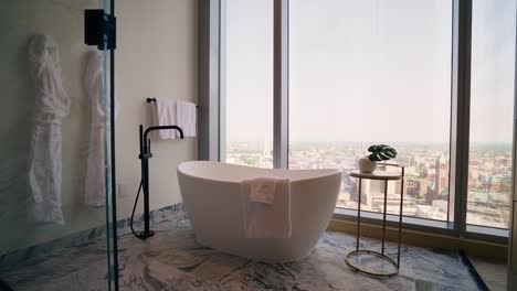 wide shot of modern white free standing bath tub in high rise bathroom with large windows and a city view