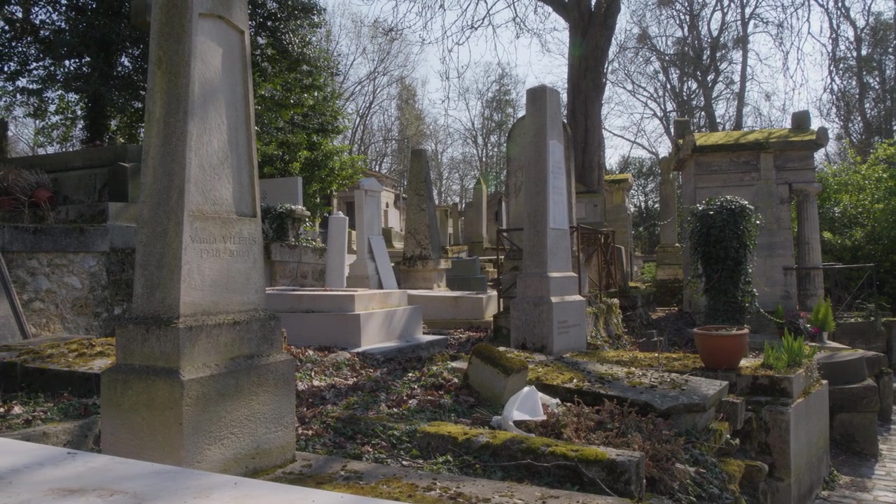 Panoramica a sinistra attraverso il Cimitero di Père Lachaise a Parigi, che mostra vecchie tombe, lapidi in pietra ricoperte di muschio, alberi e sentieri tranquilli in una luce diurna soffusa.