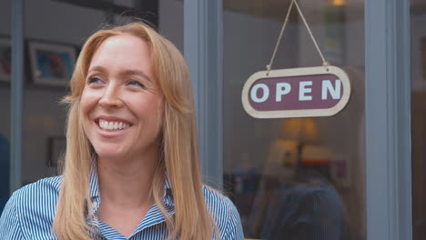 Female-Owner-Or-Staff-Outside-Shop-Or-Cafe-Standing-By-Open-Sign-In-Doorway