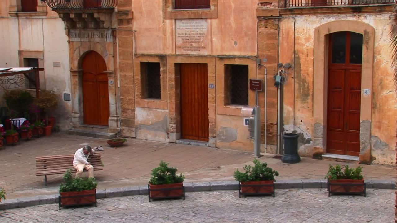 мужчина сидит на скамейке рядом с кирпичными зданиями в непосредственной близости друг от друга в цефалу италия
