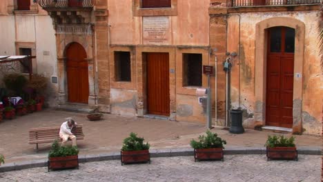 a man sits on a bench next to brick buildings within close proximity of one another in cefalu italy