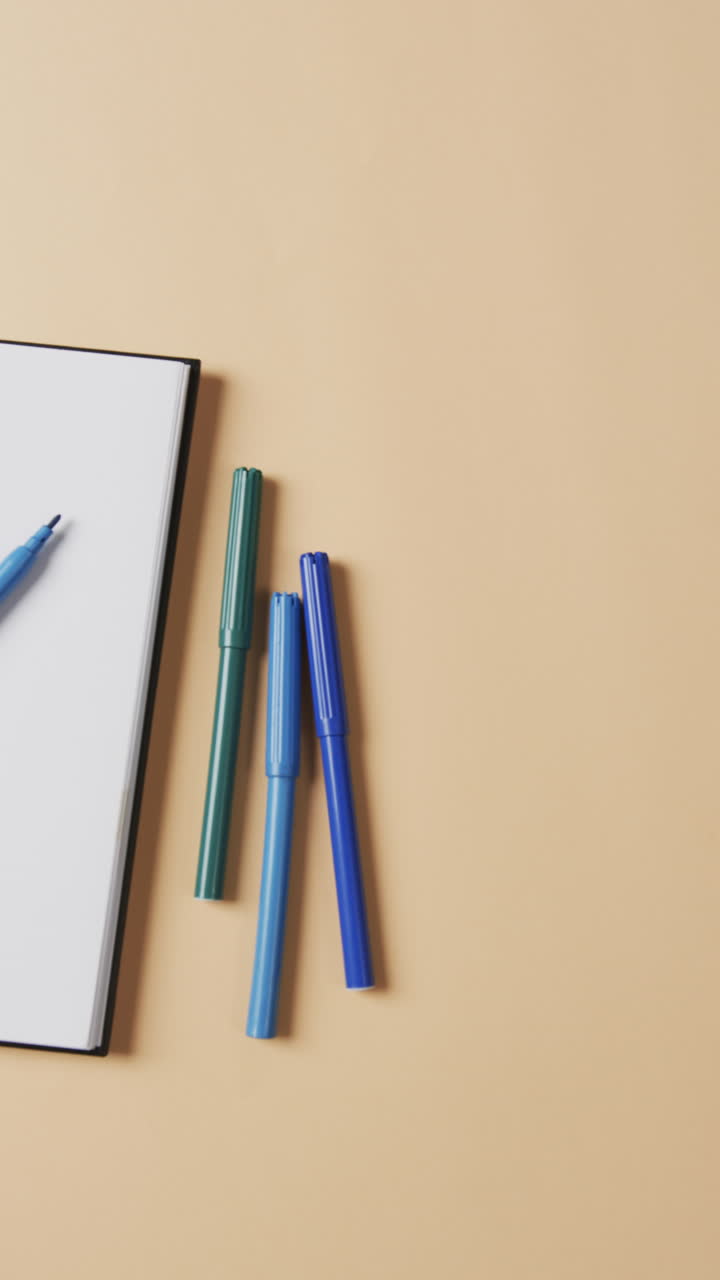 Vertical video of overhead view of notebook with blue markers on beige background, in slow motion