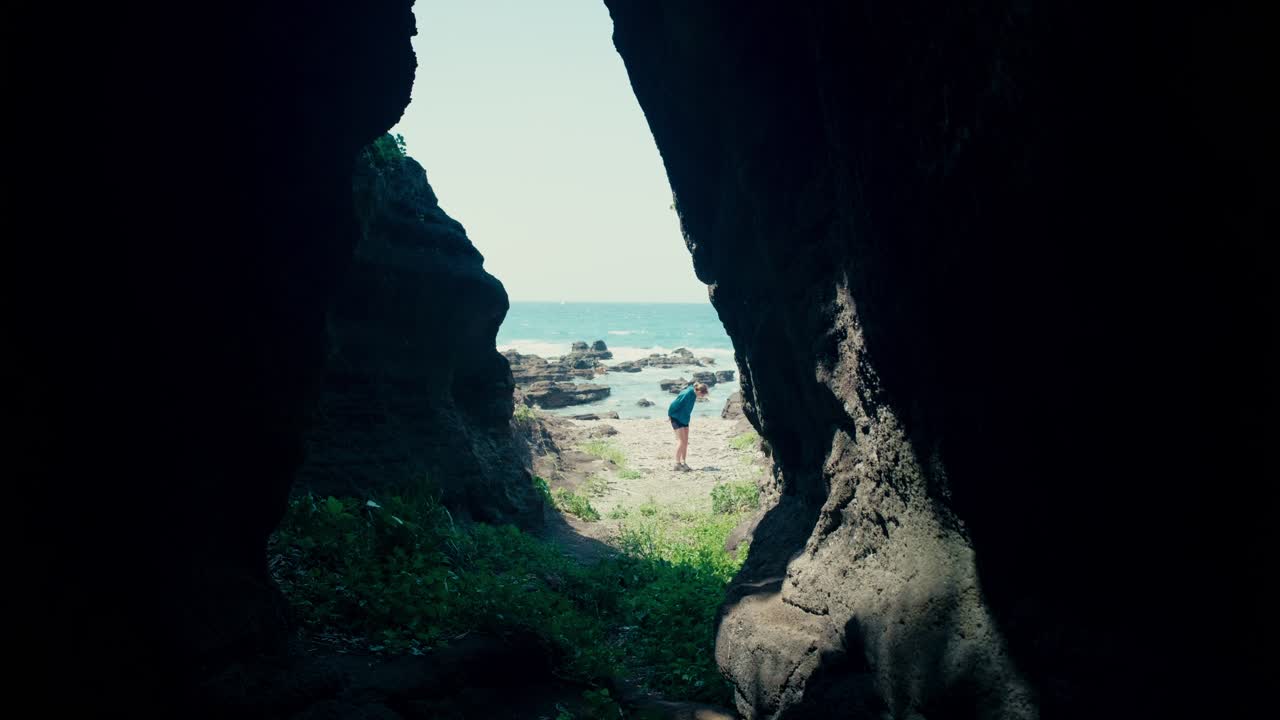 Girl walks along beach collecting shells, framed by the entrance of a seaside cave.