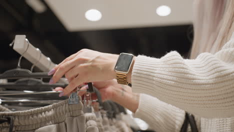 modern mother hands in knit sweater and gold smartwatch slide hangers on rack, pulling beige jogger trousers forward, testing stretch and size tag under store light, blurred boutique background