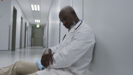 Tired-african-american-male-doctor-sitting-on-hospital-corridor-holding-face-mask