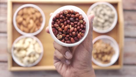 Hand holding a bowl of peanuts with other nuts on a wooden tray