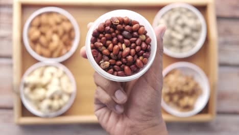 hand holding a bowl of peanuts with other nuts on a wooden tray