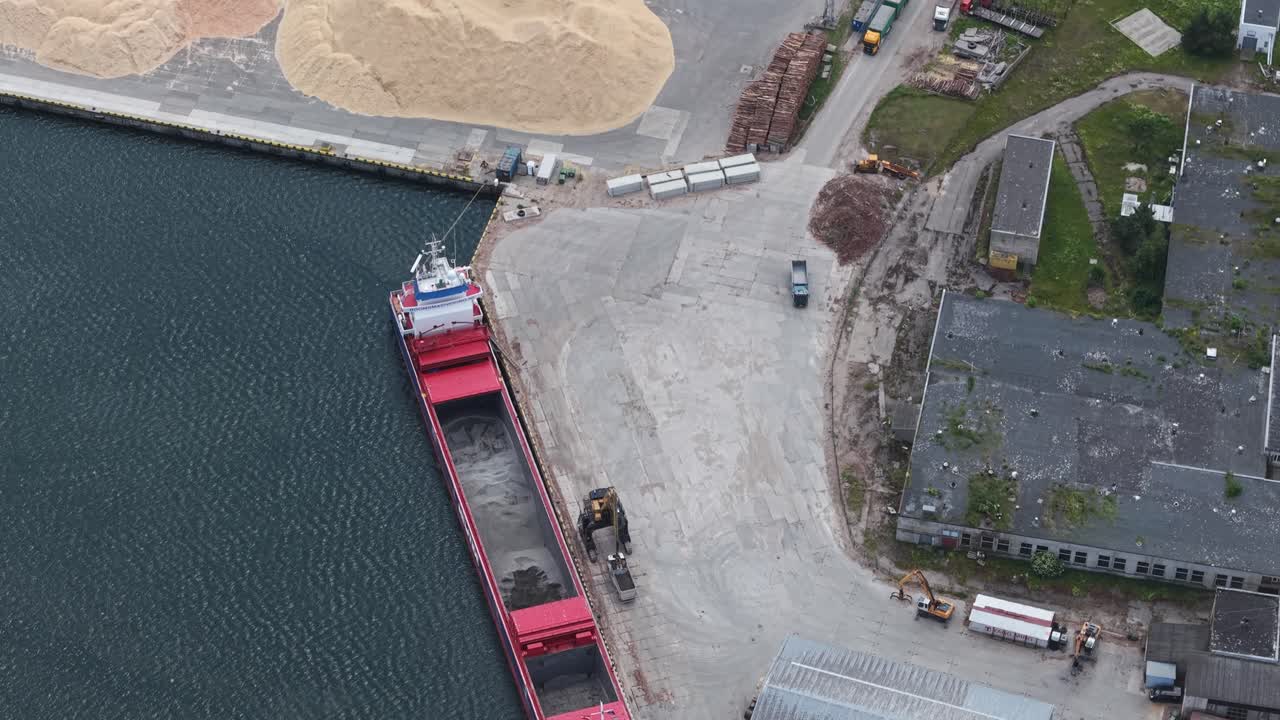 Vista aerea dall'alto di una nave da carico attraccata in un porto industriale con container e banchina visibili; un escavatore carica la nave