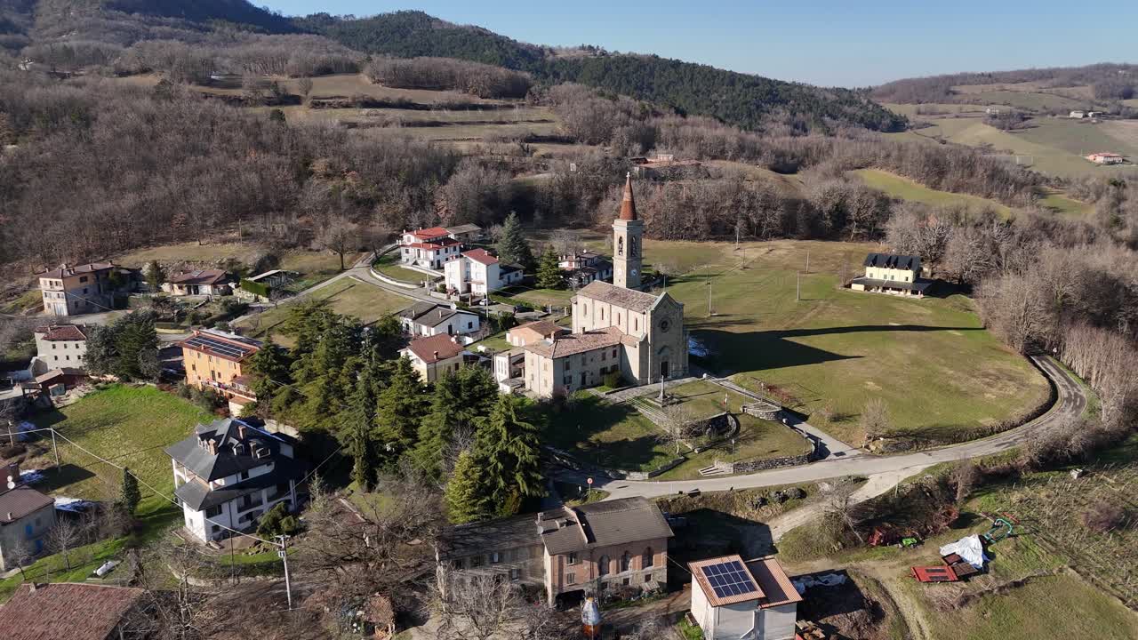 La Chiesa dei Santi Salvatore e Gallo si trova a I Rabbini, Morfasso, con la sua facciata in pietra e il campanile incorniciati da case del villaggio, cipressi e i dolci pendii degli Appennini.