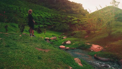 a young woman is standing by a flowing stream near a tea plantation field at cameron highlands in malaysia