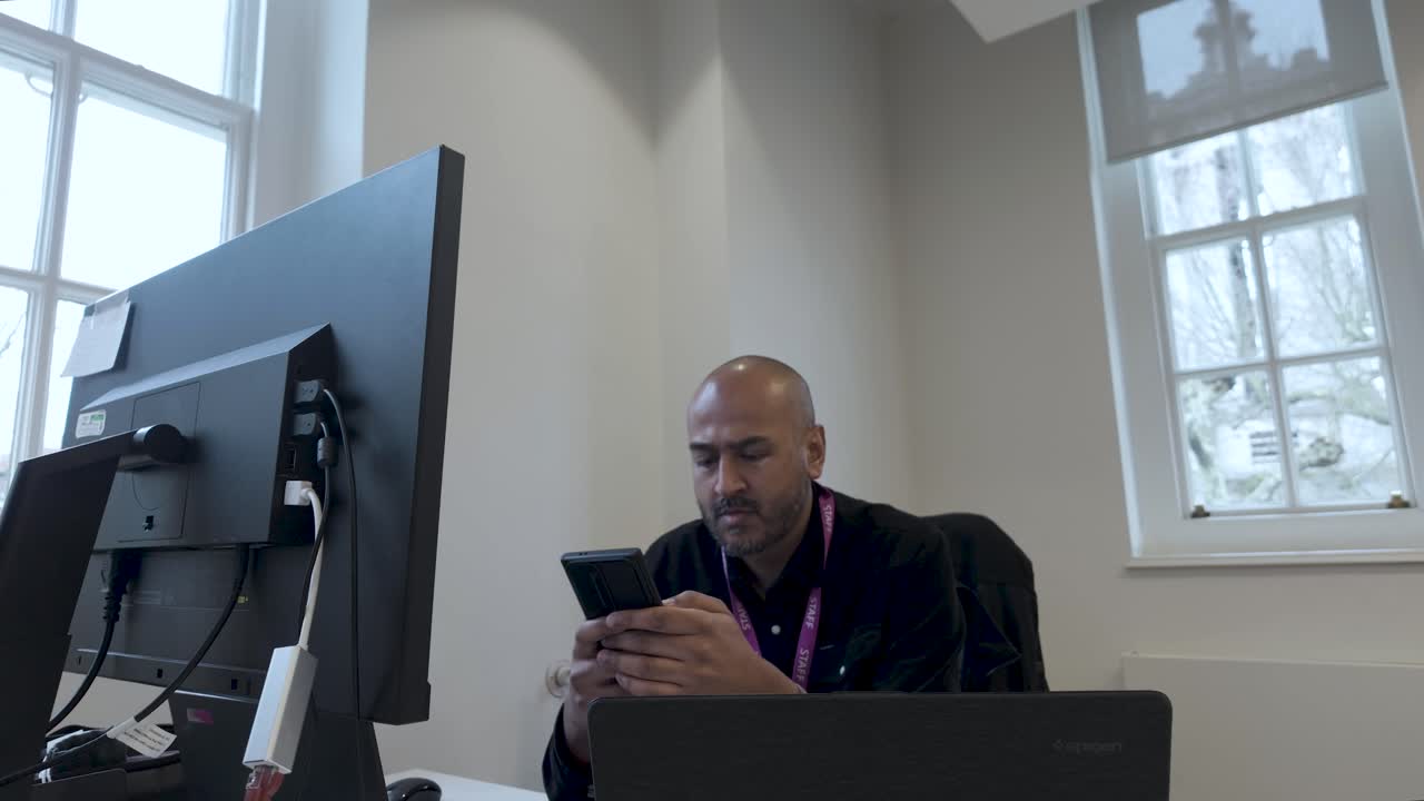 South Asian British Bald Male Office Worker Using Mobile Phone In Hands Sitting At Desk In Business Setting