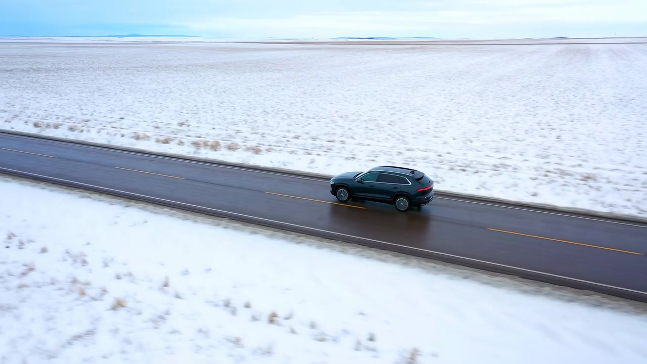 SUV scuro che guida da sinistra a destra lungo una strada rurale bagnata a due corsie, delimitata da campi innevati, campagna invernale aperta, cielo coperto, asfalto riflettente, una scena di viaggio isolata con luce naturale calma.