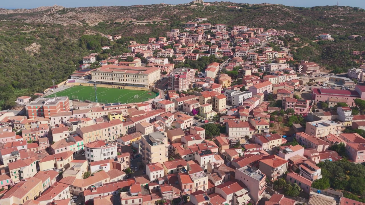 Veduta aerea ascendente di tetti in terracotta e campo da calcio a La Maddalena, una popolare destinazione per la vela e i viaggi con viste mozzafiato, storia e paesaggio costiero mediterraneo in Sardegna, Italia