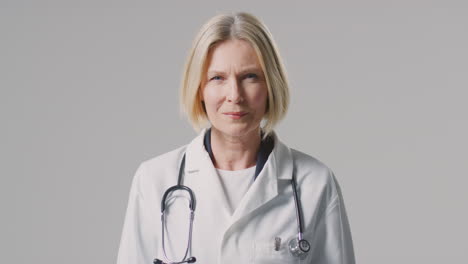 studio portrait of mature female doctor with stethoscope wearing white coat against plain background