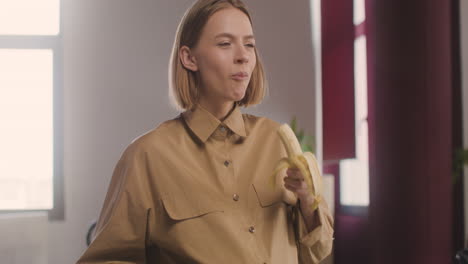 happy pregnant woman standing eating a banana and looking at camera in the office while caressing her belly