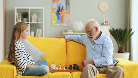 Un-Anciano-Con-El-Pelo-Gris-Jugando-Al-Ajedrez-Con-Su-Linda-Nieta-Adolescente-En-El-Sofá-De-Casa