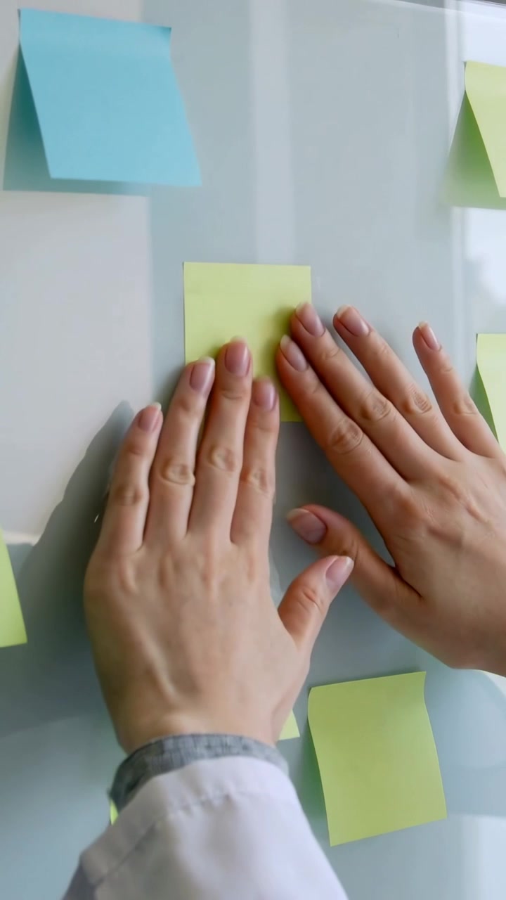 Video verticale: Mani in maniche che posizionano e lisciano un appunto verde su una lavagna di vetro, pianificando.