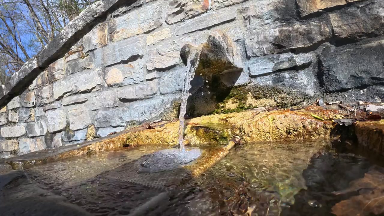 Acqua dolce limpida emerge da un piccolo sbocco di sorgente in un muro di contenimento in pietra, scorrendo dolcemente attraverso un canale poco profondo circondato da muschio e texture di rocce bagnate.