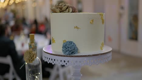 Wedding-Day-stock-clip-of-wedding-cake-single-tier-simple-with-gold-flakes-and-hand-made-wood-flowers-with-reception-people-in-background