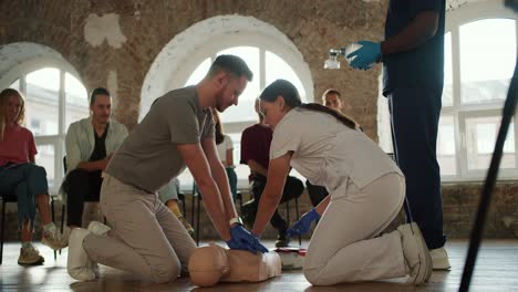 Close-up-shooting:-a-bearded-brunette-man-in-a-gray-T-shirt-with-a-nurse-girl-in-a-white-uniform-demonstrates-the-rules-of-first-aid-with-a-mannequin-in-front-of-the-public-against-the-background-of-brick-walls-and-large-windows