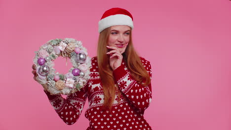 Adult-smiling-girl-in-red-Christmas-holiday-sweater-hold-raise-point-finger-on-festive-wreath-toy