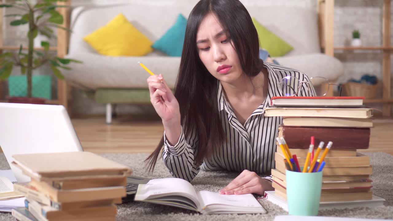 リビングの床にノートパソコンと本を広げて横たわり、試験勉強をしている疲れたアジア人学生。