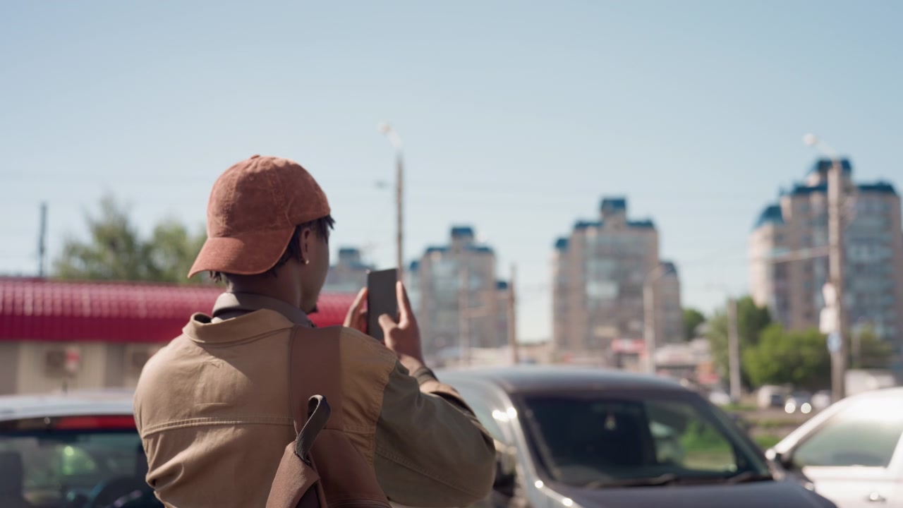 Hombre con gorra marrón y bolso toma fotos con su smartphone en un estacionamiento urbano, capturando edificios altos bajo un cielo azul claro. Hay coches estacionados cerca, mostrando una escena al aire libre.